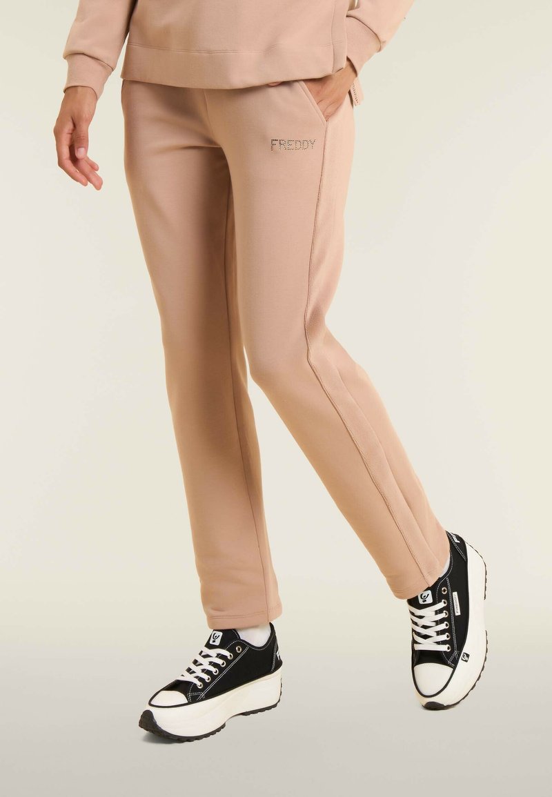 Pantalones de chándal beige en tejido suave con corte slim. Presentan un logotipo bordado de "FREDDY" y bolsillos laterales. Zapatillas de plataforma en negro y blanco.