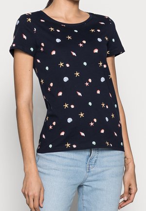 Femme portant une chemise à manches courtes bleu marine avec des imprimés multicolores de coquillages et étoiles de mer et un jean bleu clair.
