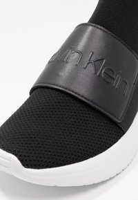 Černá atletická bota s síťovinovým svrškem, která má lesklý černý kožený pásek s embosovaným nápisem "Calvin Klein" a bílou gumovou podešví.