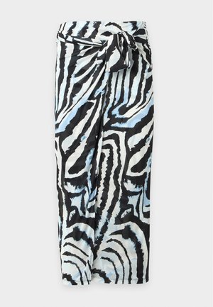 Donna che indossa un vestito con stampa a zebra nero, bianco e blu, che tiene una borsa nera e indossa mule nere con tacco su sfondo semplice.