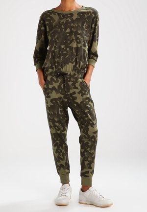 Personne portant un sweat-shirt et un jogging assortis vert olive et noir à motif abstrait, avec des baskets blanches, debout les mains dans les poches.