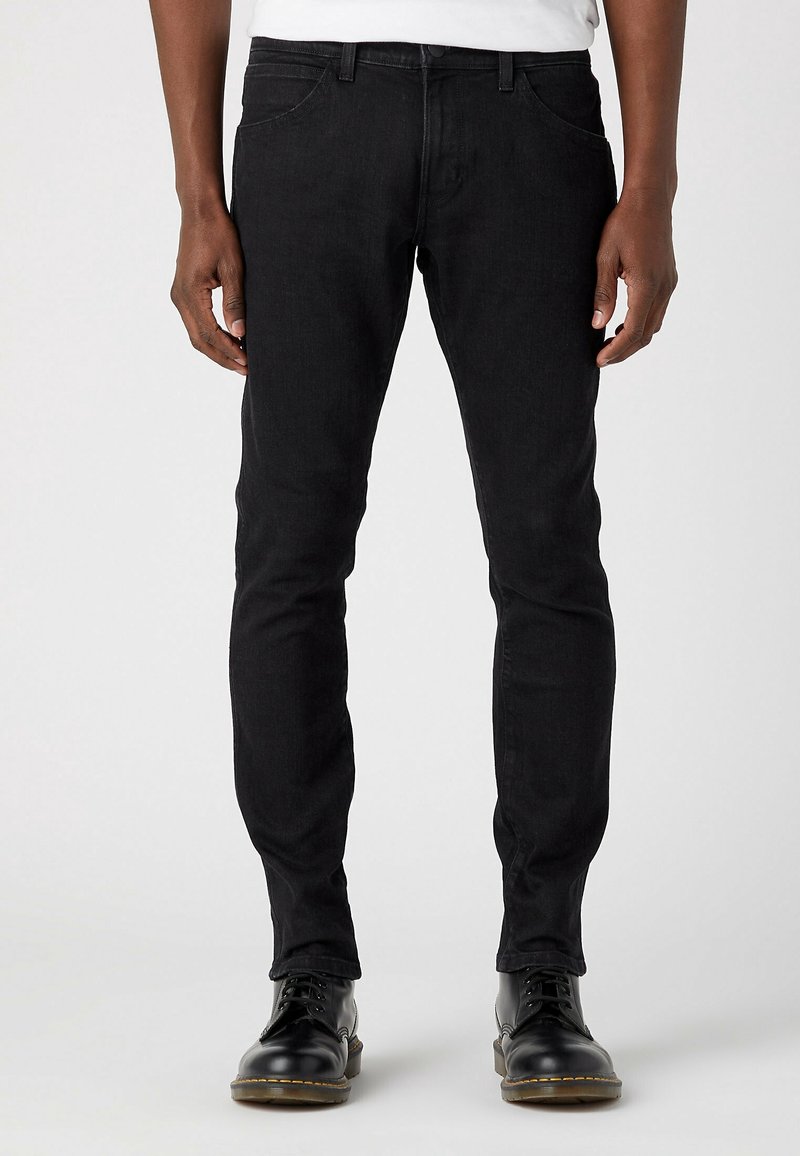 Personne portant un jean noir slim et des bottes noires à lacets, debout devant un fond clair uni.