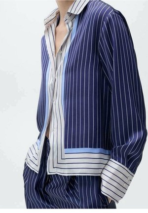 Personne portant une chemise rayée bleu marine et blanche et un pantalon assorti, les mains dans les poches et le col ouvert.