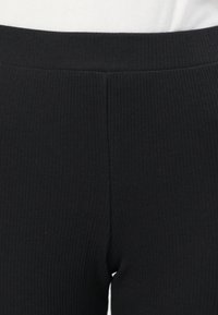 Gros plan de la ceinture et de la partie supérieure de la cuisse de leggings en tissu côtelé noir portés par une personne avec un haut blanc.