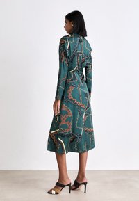 Grünes Midikleid mit langen Ärmeln, das ein Kettenmuster in Gold, Schwarz und Braun zeigt. Glattes Gewebe und fließende Silhouette. Schwarze Absatzsandalen.