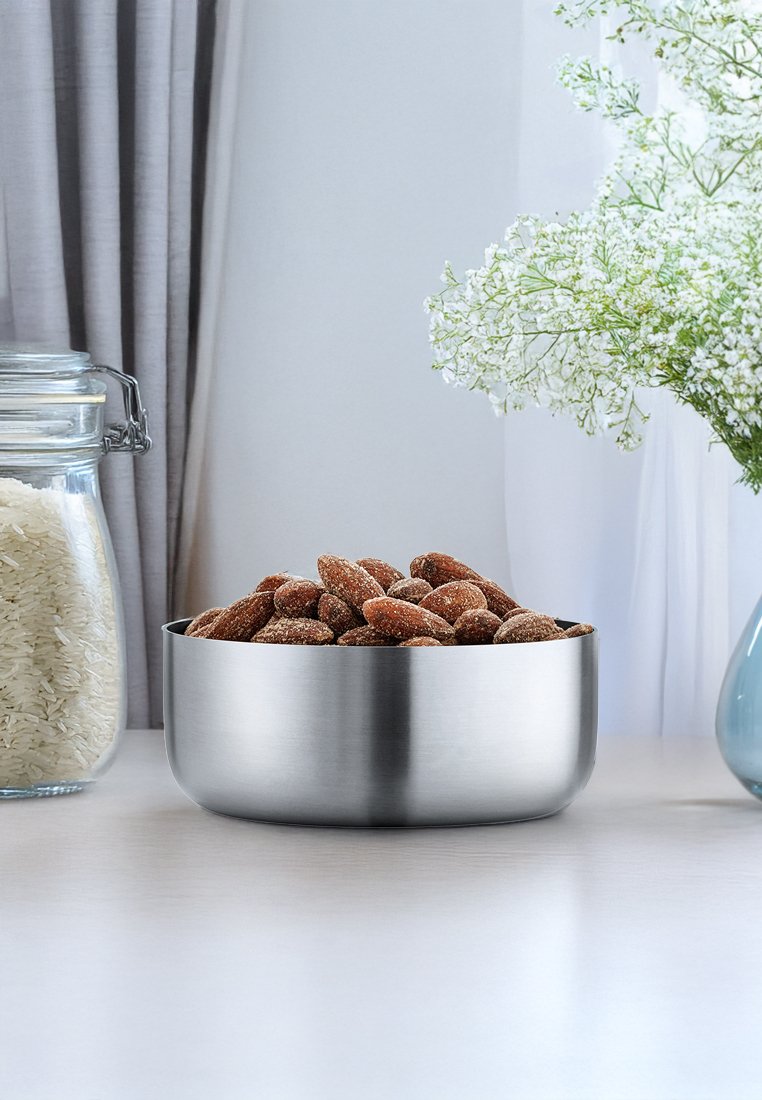 Edelstahl-Schüssel gefüllt mit braun beschichteten Mandeln, auf einer hellen Oberfläche platziert, in der Nähe eines Glasgefäßs mit Reis und einer blauen Vase mit weißen Blumen.