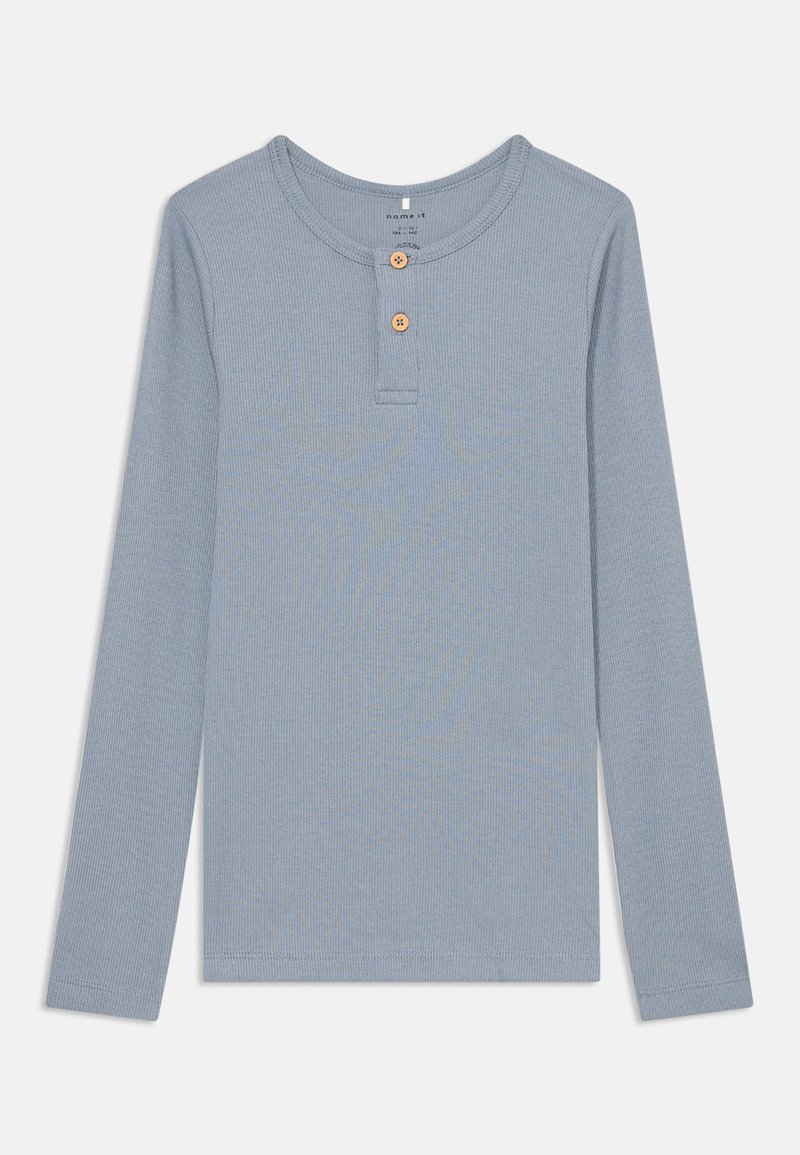 Ilgas rankovėmis šviesiai mėlynas raukšlėtas marškinėliais su apvaliu iškirpimu ir dviem mediniais mygtukais priekyje. Paprastas ir tekstūruotas dizainas.