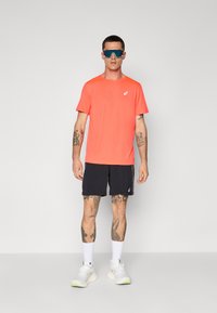 Sporty outfit med en lys koralfarvet T-shirt med korte ærmer, sorte shorts, hvide sneakers og tonede solbriller. Tatoveringer synlige på armene.
