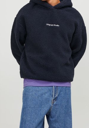 Person wearing black fleece hoodie with "Originals Studio" text, purple shirt underneath, and blue jeans, standing against plain background.