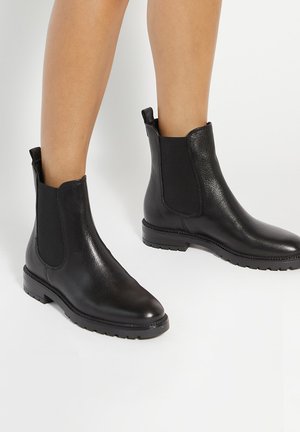 Bottes courtes en cuir noir avec panneaux latéraux élastiques portées sur des jambes nues sur fond blanc.