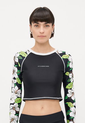 Woman wearing a black crop top with floral-patterned long sleeves and "IT'S NOW COOL" text on the chest against a plain light background.