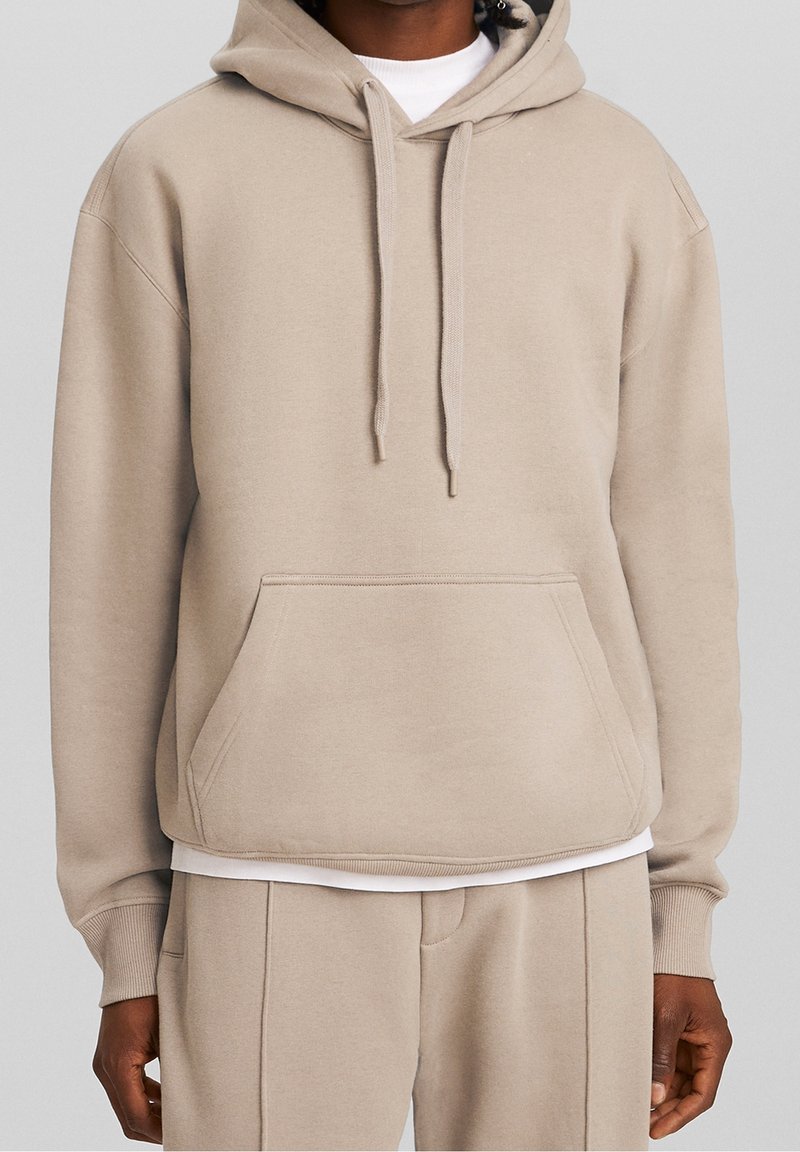 Felpa beige con cappuccio in tessuto morbido, dotata di tasca a marsupio, polsini e orlo a costine, con una vestibilità rilassata. Indossata sopra una camicia bianca.