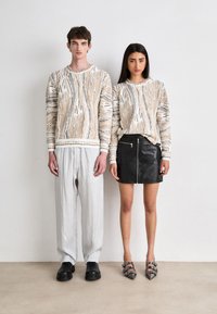 Două figuri stau una lângă cealaltă. Una poartă un pulover bej cu un model și pantaloni gri; cealaltă poartă o fustă neagră cu fermoar și pantofi argintii.