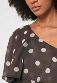 Γυναίκα με σγουρά μαλλιά που φοράει μαύρο φόρεμα με έναν ώμο έξω και μεγάλα λευκά πουά, εμφανίζοντας τα χείλη και το άνω μέρος του στήθους.