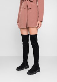 Bottes en daim noir au-dessus du genou avec un bout arrondi, une finition texturée et une semelle épaisse. Elles sont dotées d'une fermeture éclair latérale pour un enfilage facile.