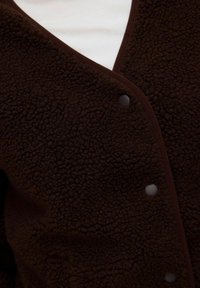 Veste en polaire texturée brun foncé avec des boutons-pression portée sur un haut blanc, montrant un gros plan de la zone de fermeture avant.