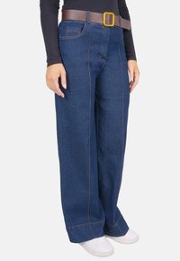 Pantalon en denim à jambes larges de couleur bleu foncé, avec des coutures orange et une ceinture marron avec une boucle jaune. Associé à des baskets blanches.