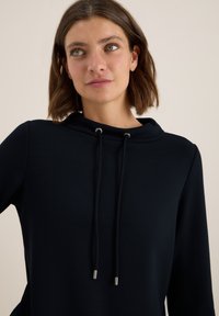 Navyblauer Hoodie, weicher Stoff, mit hohem Kragen, silbernen Ösen und Kordeln mit Metallenden. Lässiger Schnitt und glatte Oberfläche.