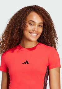 Crvena atletska majica napravljena od glatkog materijala, sa rebrastim okruglim izrezom, kratkim rukavima i crnim bočnim trakama s Adidas logotipom.