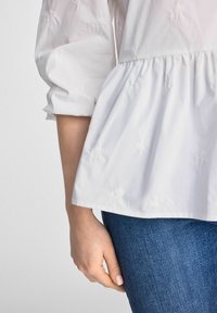 Biała bluzka z teksturowanym haftem kwiatowym, rękawami w kształcie dzwonu i talią peplum, zestawiona z ciemnoniebieskimi dżinsami.