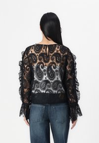 Schwarze Spitzenbluse mit Paisleymuster, Rüschenärmeln und einem Schlüssellochdetail auf der Rückseite. Kombiniert mit blauen Jeans.