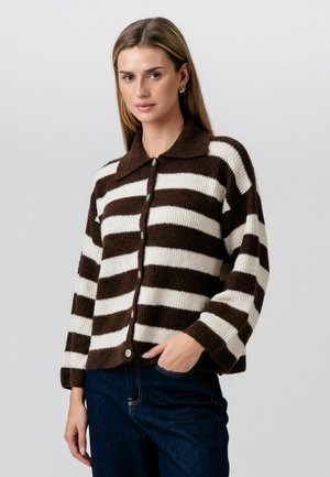 Kobieta ubrana w brązowo-biały sweter w poziome paski zapinany na guziki, z długimi rękawami i kołnierzem, w zestawieniu z ciemnoniebieskimi dżinsami.