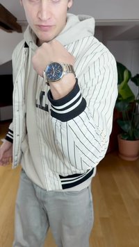 Sidabrinis laikrodis su mėlynu ciferblatu, metaliniu apyranke ir kontrastingais valandų žymekliais. Nešiojamas ant riešo, derinamas su dryžuota švarku ir smėlio spalvos vaisiu.