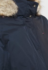 Námořnicky modrá bunda s kapucí olemovanou umělým kožíškem, vybavená patentními knoflíky a s logem jako detailem. Materiál působí hladce s jemným leskem.