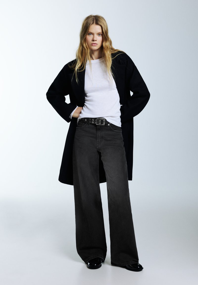 Čierny kabát cez biele tričko, skombinovaný so širokými sivými džínsami a čiernym opaskom so striebornými ozdobami. Lesklé čierne topánky dotvárajú vzhľad.