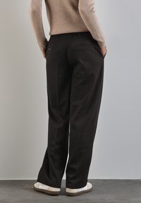 Pantaloni neri a vita alta con una vestibilità rilassata, dotati di tasche laterali, in un tessuto testurizzato e abbinati a sneakers bianche su uno sfondo grigio.