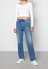 Vit långärmad croppad t-shirt med en röd broderad logga, ihopparad med ljusblå denimjeans med lös passform och bleknade områden. Vita sneakers.