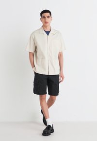 Camisa de manga corta beige con botones y dos bolsillos frontales, combinada con pantalones cortos cargo negros. El modelo lleva zapatos negros y calcetines blancos.