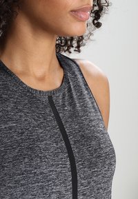 Femme aux cheveux bouclés portant un haut de sport sans manches gris foncé avec une bande noire verticale à l'avant, visible des épaules jusqu'aux lèvres.