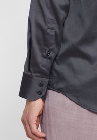 Camicia nera a maniche lunghe con texture e bottoni ai polsini, indossata con pantaloni chiari. Il tessuto appare liscio con una leggera lucentezza.