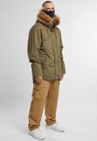Olivno zelena parka z odstranljivo kapuco obloženo s krznom, sprednjimi žepi in elastičnim pasom, v kombinaciji s bež cargo hlačami in belimi športnimi čevlji.