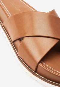 Bruine leren slip-on sandaal met een crisscross bandontwerp, witte geribbelde zool en contrasterende witte stiksels langs de randen.