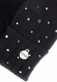 Bonnet noir en tricot orné de strass transparents éparpillés et d'une étiquette ronde blanche arborant la marque "GUESS". Texture douce et design côtelé.