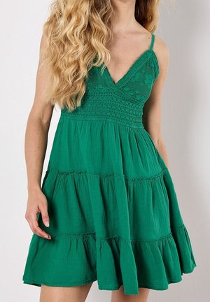 Femme portant une robe verte sans manches avec une jupe à volants et un corsage texturé, debout devant un fond clair uni.