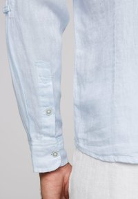 Camisa de lino azul claro con mangas largas, detalles de botones en los puños y un dobladillo ligeramente curvado. Tejido texturizado con trama visible.