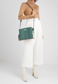 Borsa a tracolla verde con texture, base color beige chiaro, forma quadrata, tracolla regolabile, dettaglio a frange e hardware in tono oro.