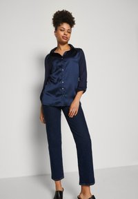 Navy Satin-Hemd mit kragen, durchgehender Knopfleiste und langen Ärmeln. Kombiniert mit dunklen Denim-Jeans und schwarzen offenen Schuhen.