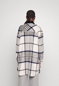 Plaid shirt jacket in cream, navy, and black. Features long sleeves, collar, button closure, and side slits. Soft, textured fabric.