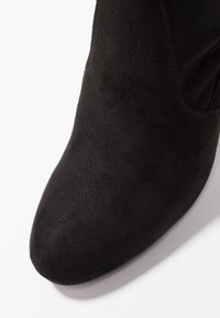 Bottine en daim noir avec un bout pointu et une texture lisse, présentant un détail de couture le long des côtés et un talon bas incurvé.