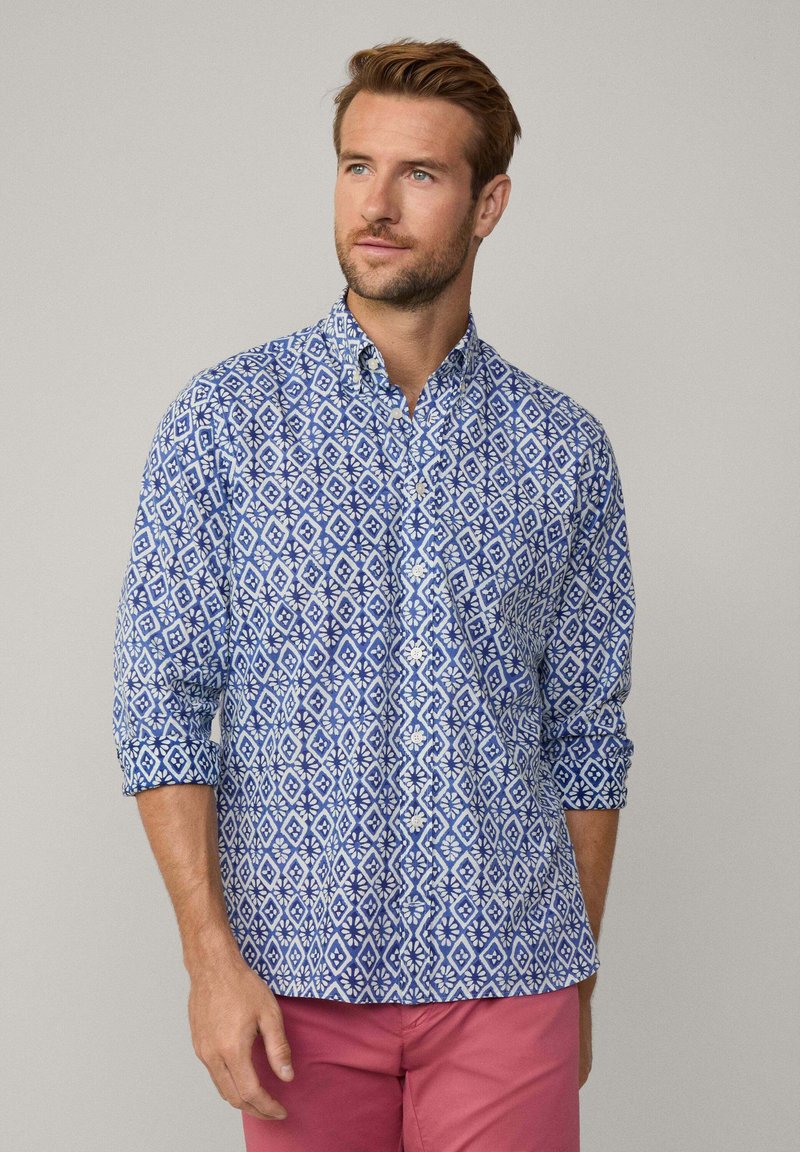 Camisa de patrón azul y blanco con diseños de diamantes y flores, con cuello abotonado y mangas enrolladas.