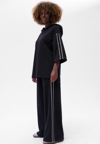 Zwarte hoodie met oversized mouwen en een kangaroo-pocket aan de voorkant, gecombineerd met bijpassende wijde broek met decoratieve witte zijstrepen.