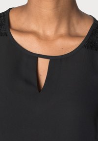 Blouse noire avec un col arrondi présentant une petite ouverture en forme de goutte, des accents noirs texturés sur les épaules et un tissu lisse.