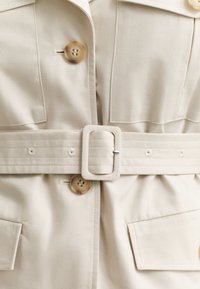 Μπεζ παλτό από ύφασμα με μεγάλες μπροστινές τσέπες, καφέ κουμπιά και φαρδύ ζωνάρι στο ίδιο χρώμα με ορθογώνια αγκράφα.