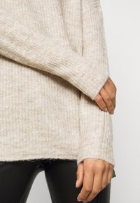 Lichtbeige gebreide trui met een gestructureerd patroon. Voorzien van lange mouwen en geribde boorden. Draagt over zwarte leren broek.