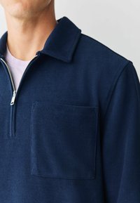Uomo che indossa un maglione zip-up a coste di colore blu navy con colletto e taschino sul petto, mentre una t-shirt bianca è visibile sotto. Sfondo grigio neutro.