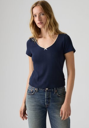 Femme aux longs cheveux blonds portant un haut bleu marine à manches courtes avec un petit nœud blanc et un jean bleu sur un fond uni.